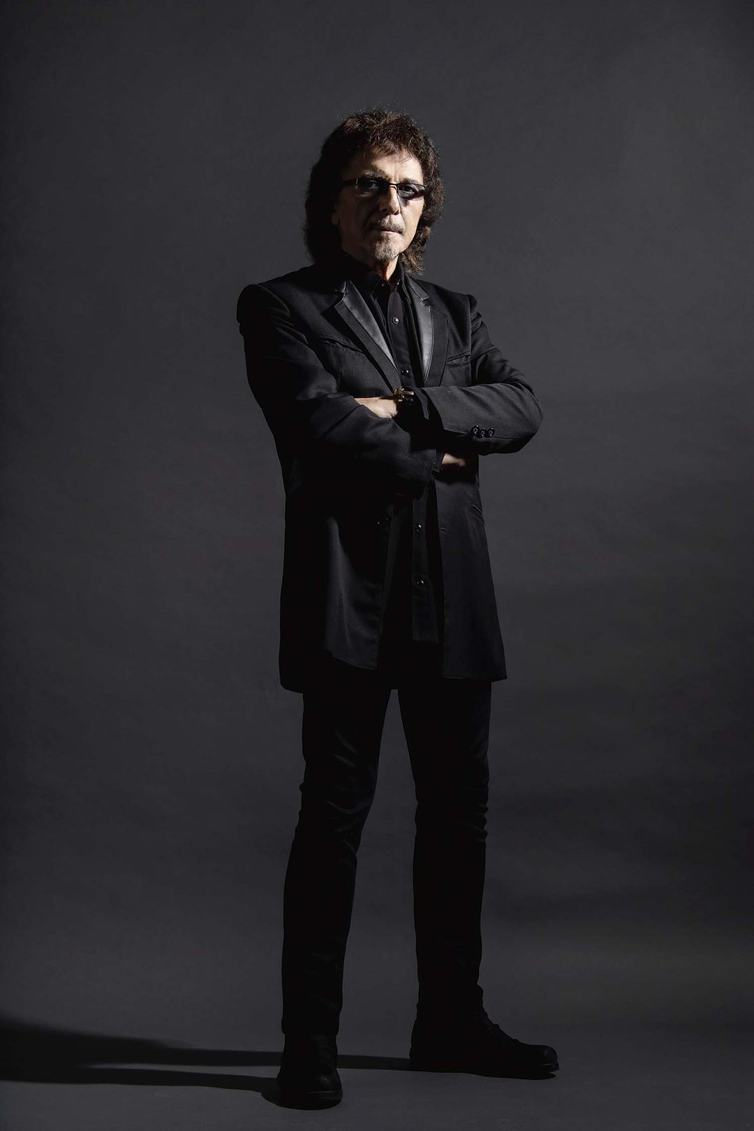 Tony Iommi con traje negro y gafas, retratado sobre fondo oscuro