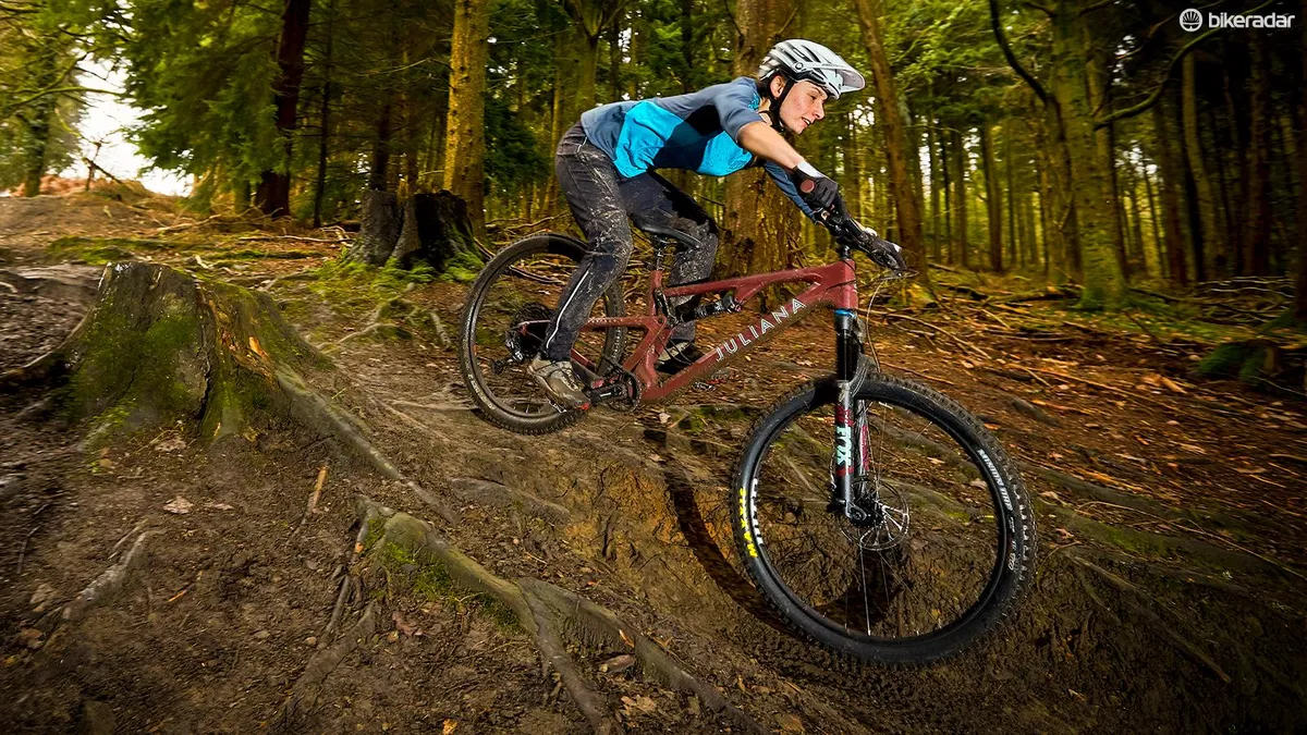 A woman in a blue top riding over roots downhill in a forest