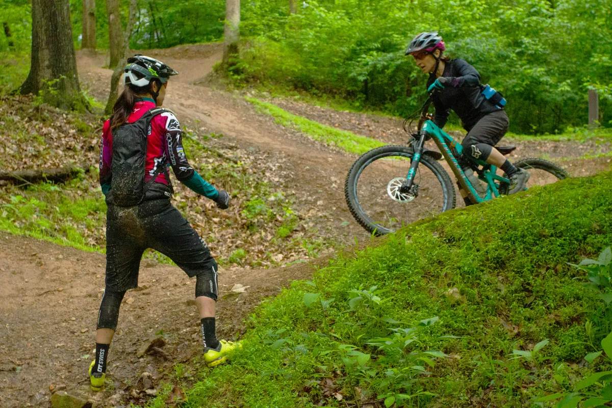 One woman mountain biker riding a trail while another woman provides coaching