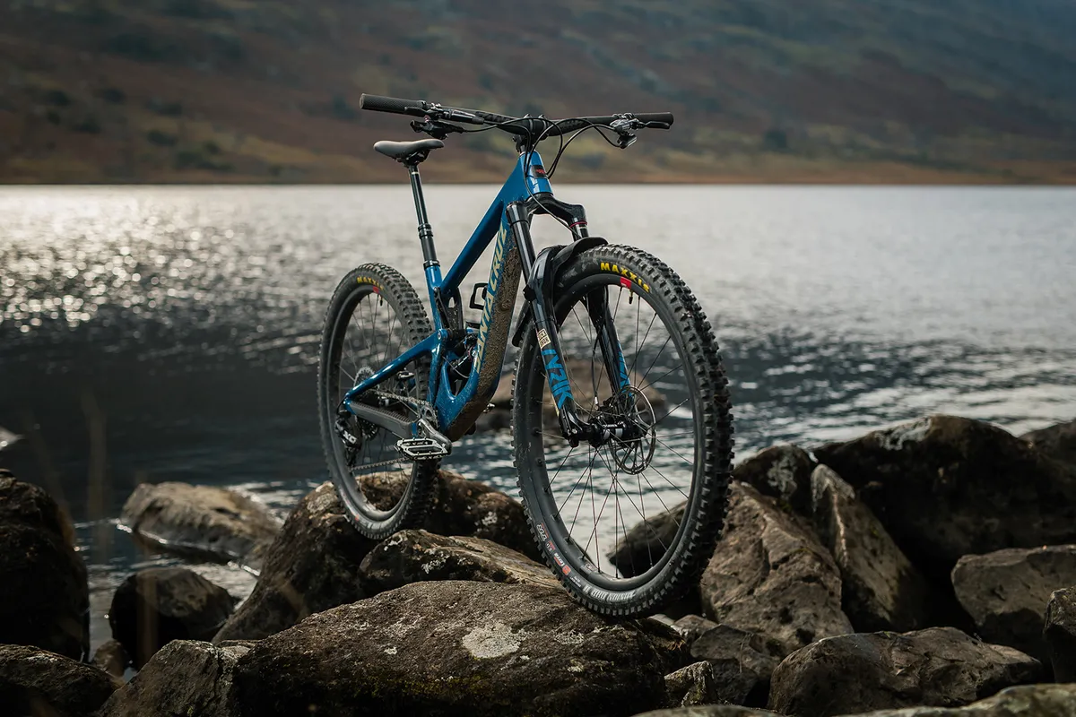 Angled pack shot of a blue full suspension mountain bike from Santa Cruz, against a backdrop of mountains and lake