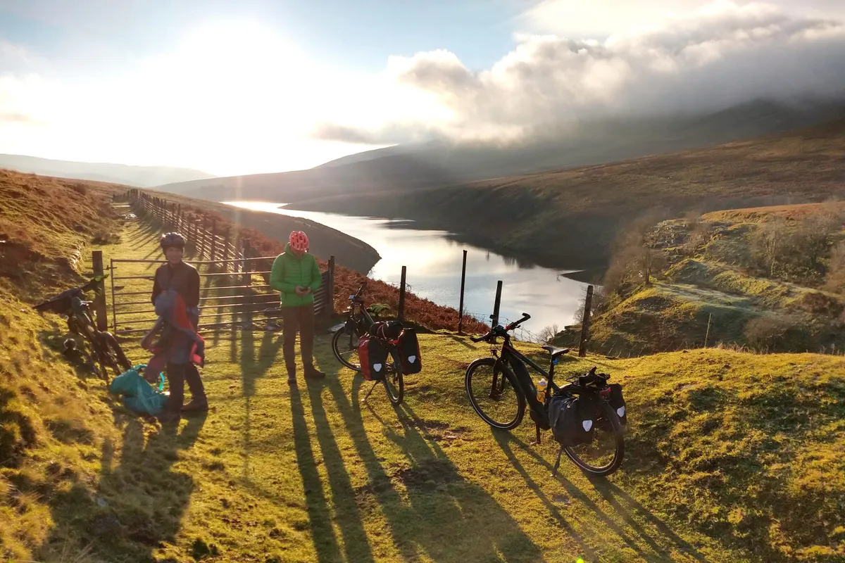 2 riders and 3 bikes on grassy track overlooking beautiful reservoir