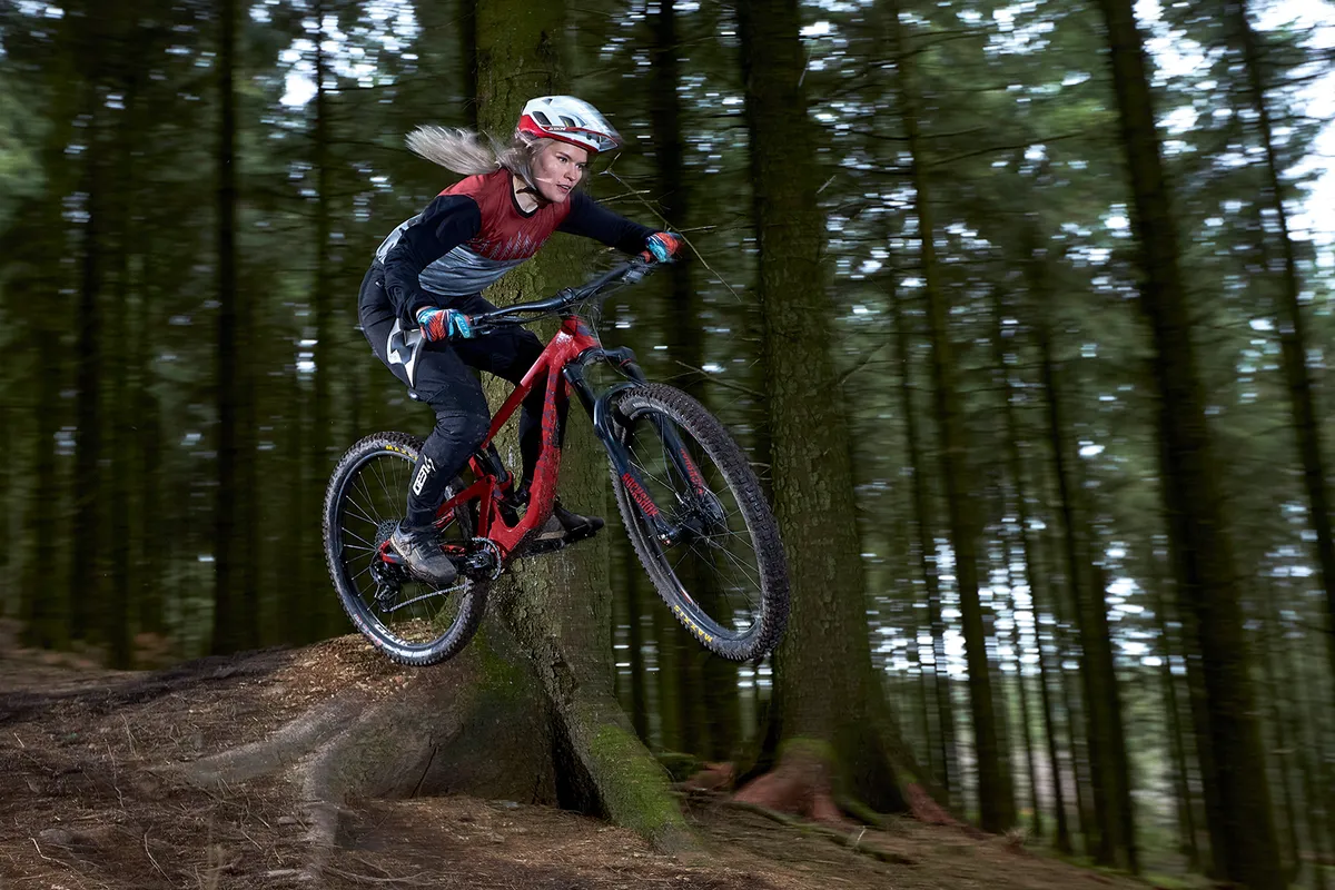 Female cyclist in red top jumping on a red full suspension mountain bike while riding through woods