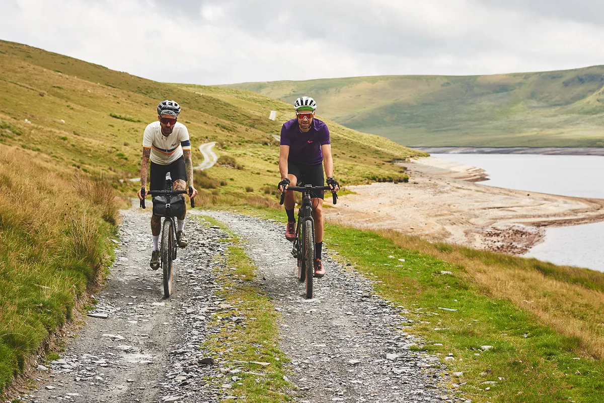 Two male cyclists riding gravel bikes next to a reservoir in Wales while on one of the Komoot routes
