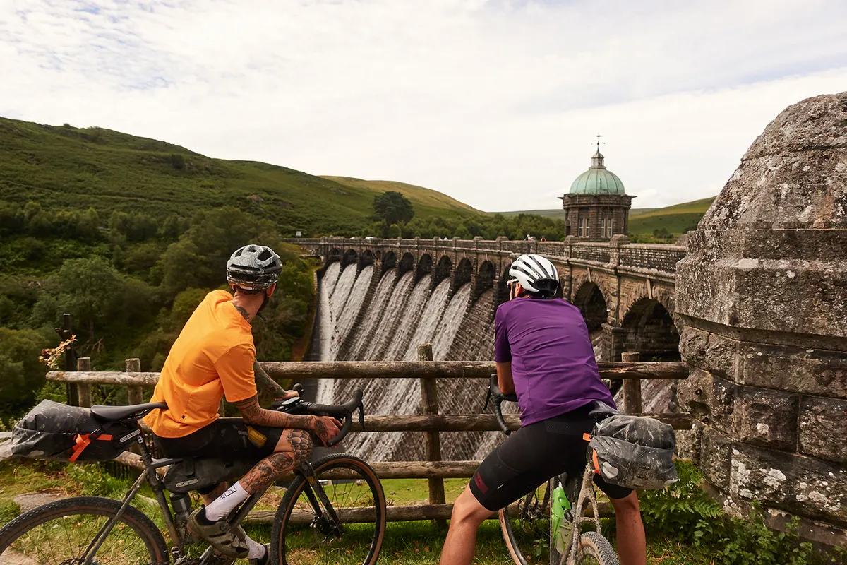 Two male cyclists riding gravel bikes next to a dam in Wales while on one of the Komoot routes