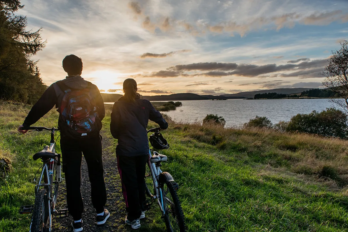 Two people holding their bikes and looking at a sunset in Carron Valley