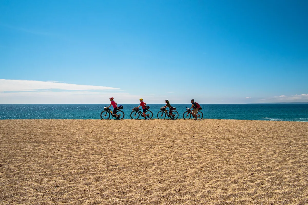 Sophie Gordon (Cycling UK), Katherine Moore (Unpaved podcast), Vedangi Kulkarni and Rob Penn (freelance journalists) push their bikes across the deep sand of the Loe Bar. The sand bar separates the Loe lagoon from the sea, and is one of several route options and shortcuts on the West Kernow Way.