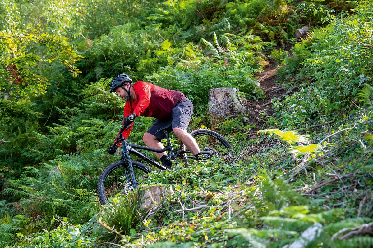 Male cyclist riding the Vitus Mythique 29 VRX full suspension mountain bike down hill through woodland
