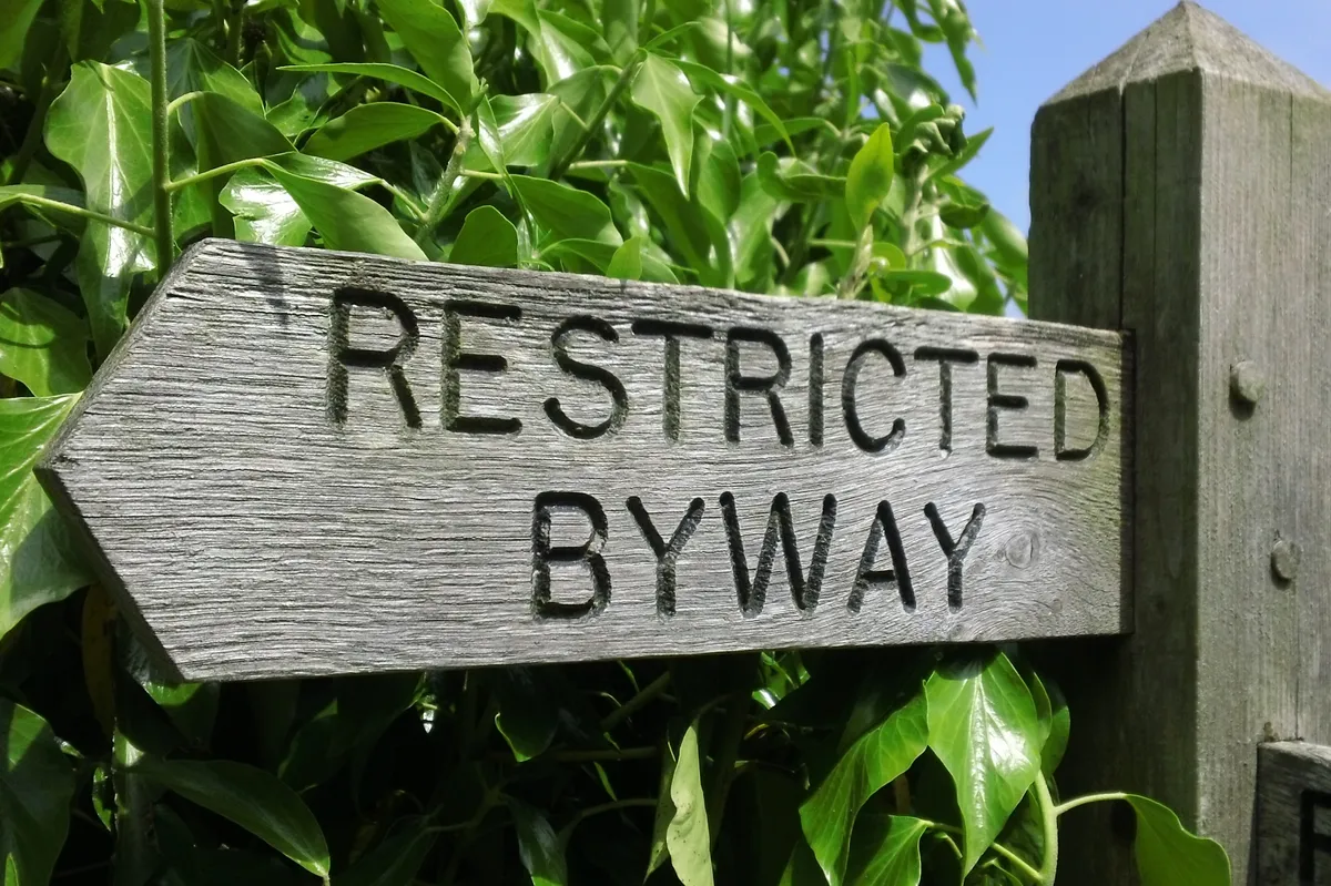 Rural 'Restricted Byway' wooden footpath sign pointing to the left. Green bush, leaves and field behind.