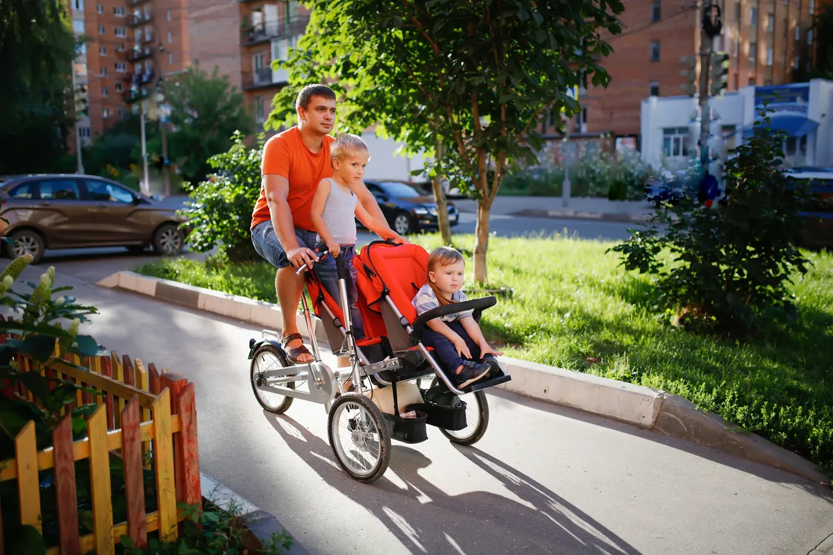 Father in an orange T-shirt rides cargo bike for two siblings of his sons in city near high-rise buildings in summer.
