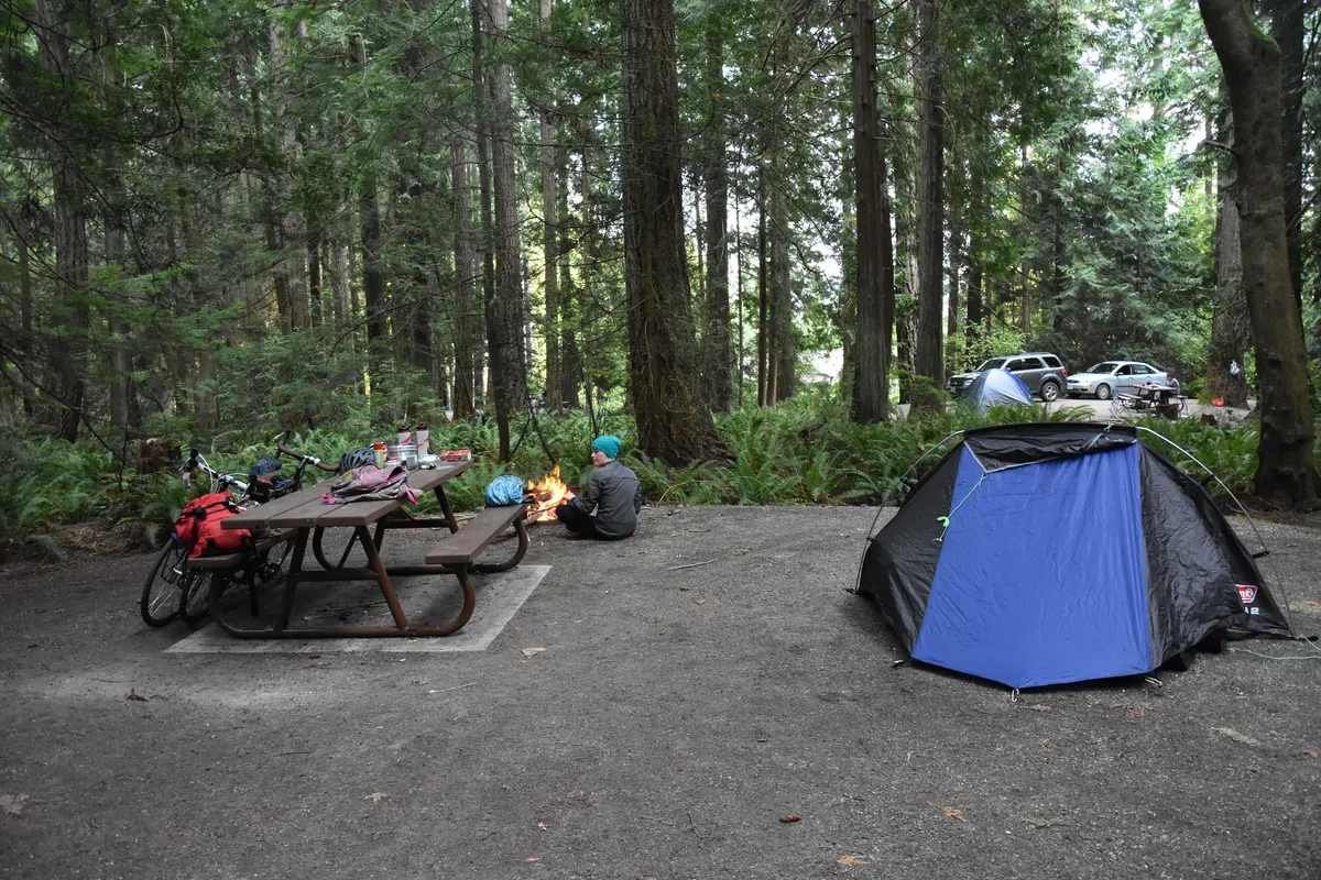 Coleman Cobra tent in woods with person tending campfire.