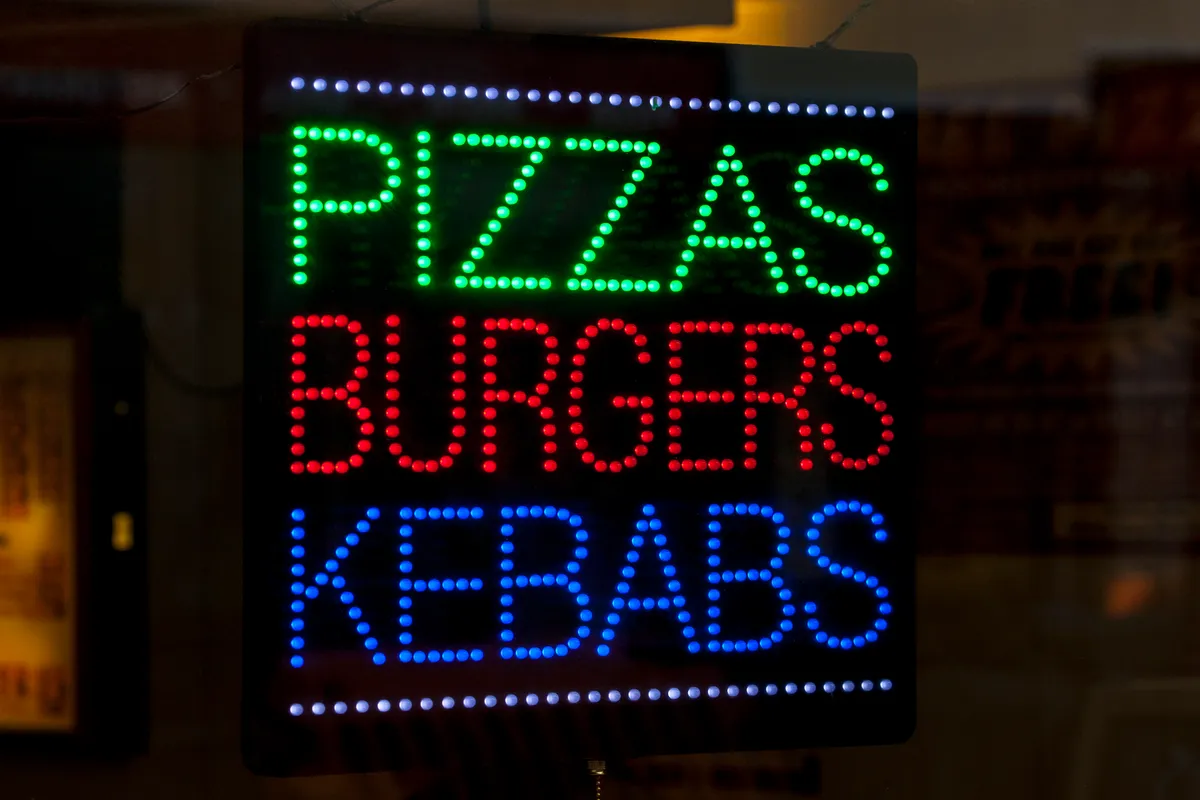 A neon sign in a takeaway shop window advertising pizzas, burgers, and kebabs