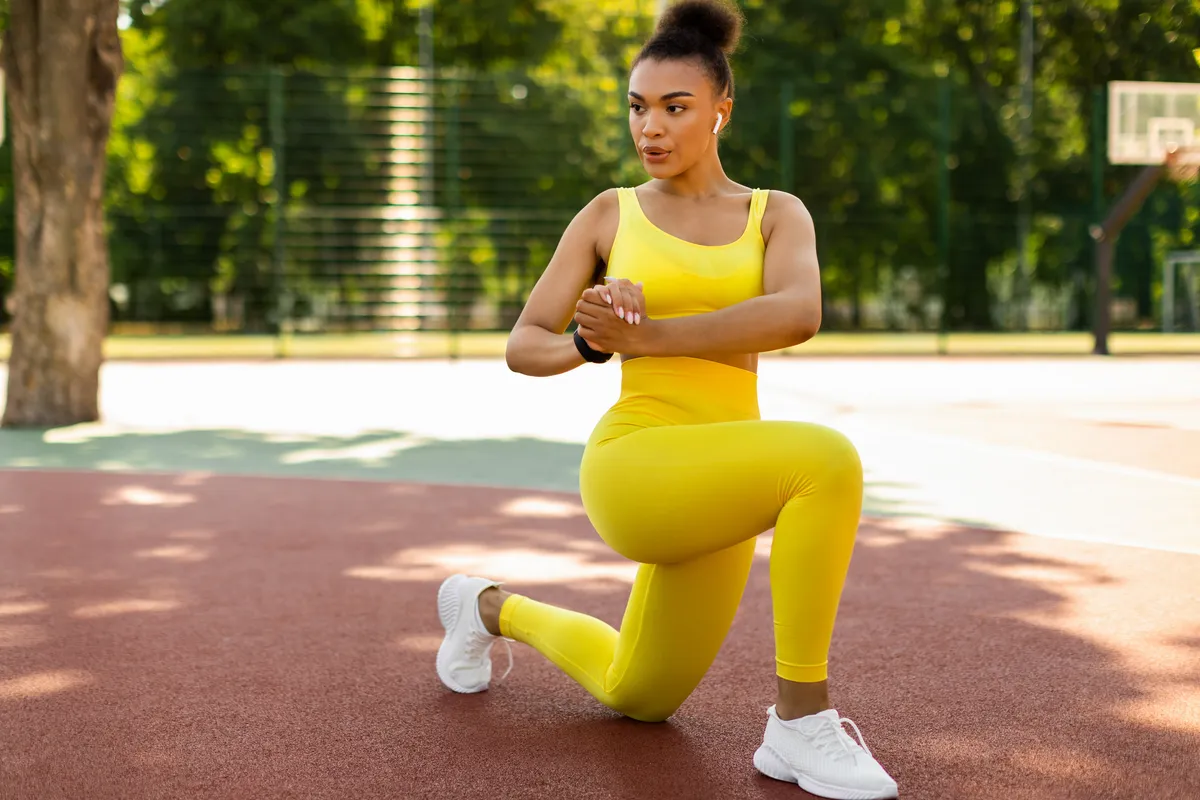 Sport And Healthy Lifestyle. Fit black woman doing curtsy lunges with twist, exercising outdoors on basketball stadium. Portrait of confident lady training in wireless earbuds, full body length