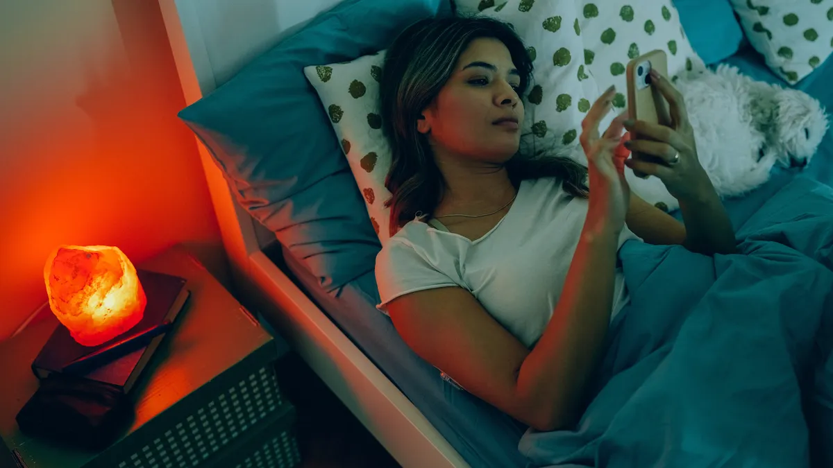 Young woman with diabetes in bed is wearing continuous glucose sensor on the back of upper arm for measuring glucose levels. She checks on mobile app whether the blood sugar is in normal range, after scanning the sensor with smartphone.