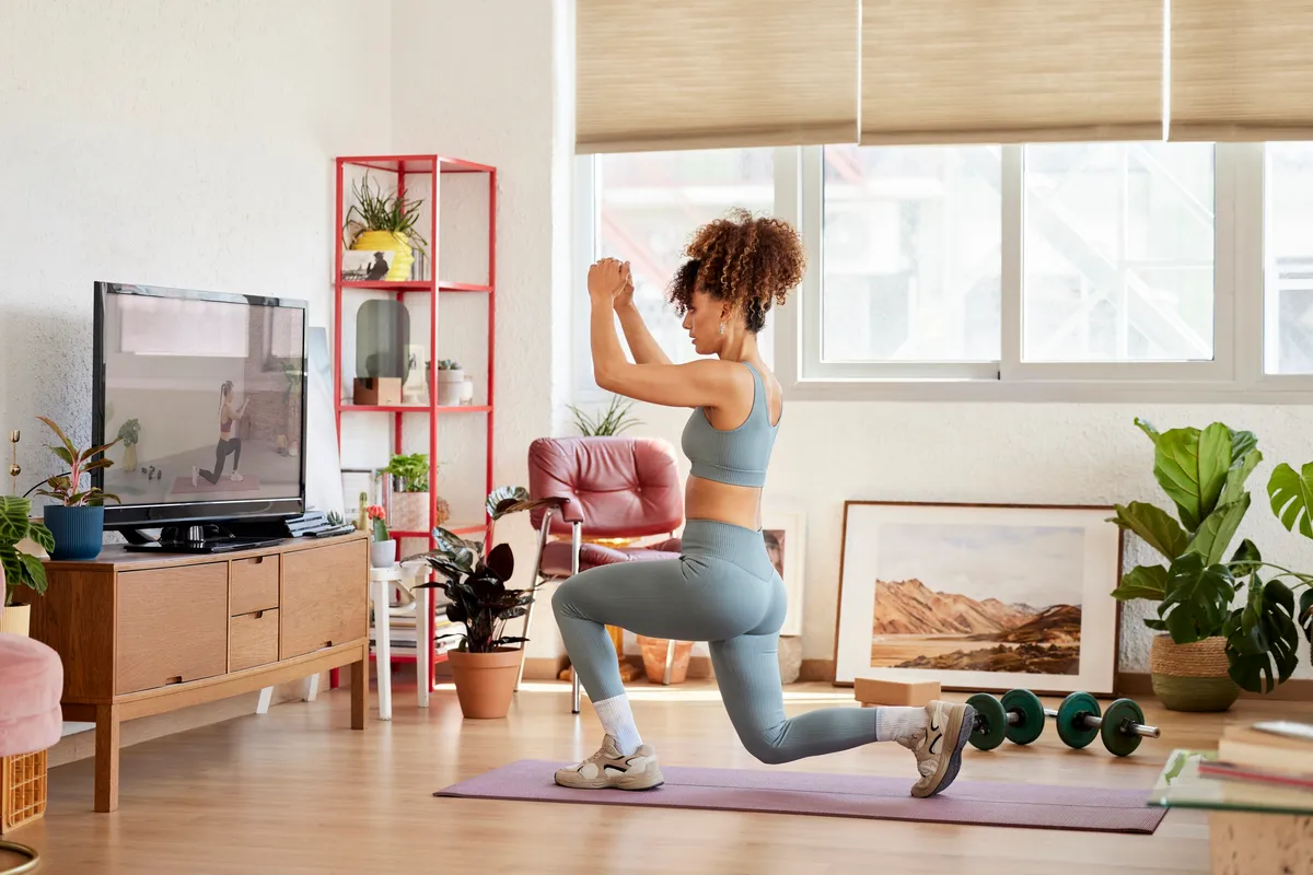 Full length of young woman learning lunges while watching online yoga class. Woman is practicing exercise at home. She is in living room.