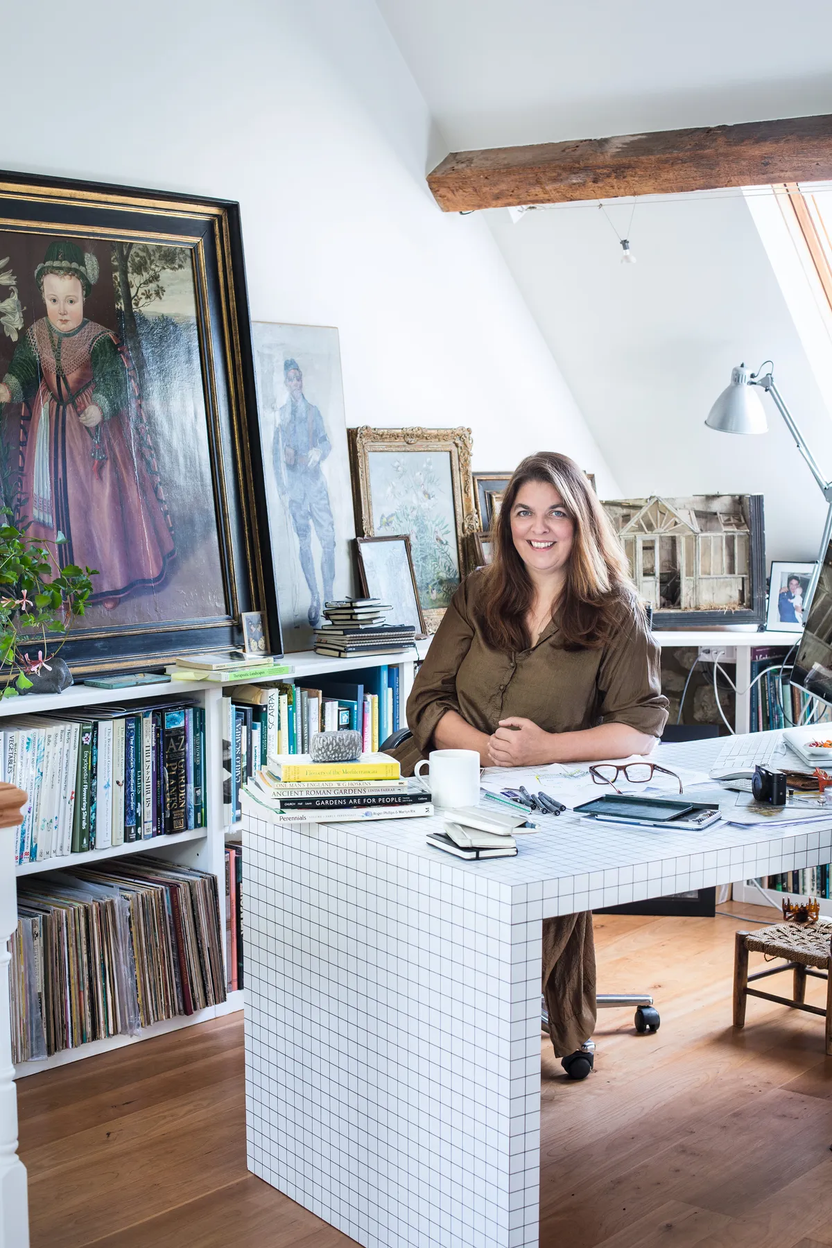 Designer Jinny Blom sits at her desk in front of a filled bookcase and antique paintings