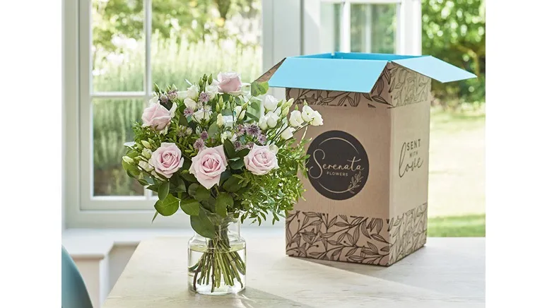 A vase of flowers in front of a cardboard box in front of a window.