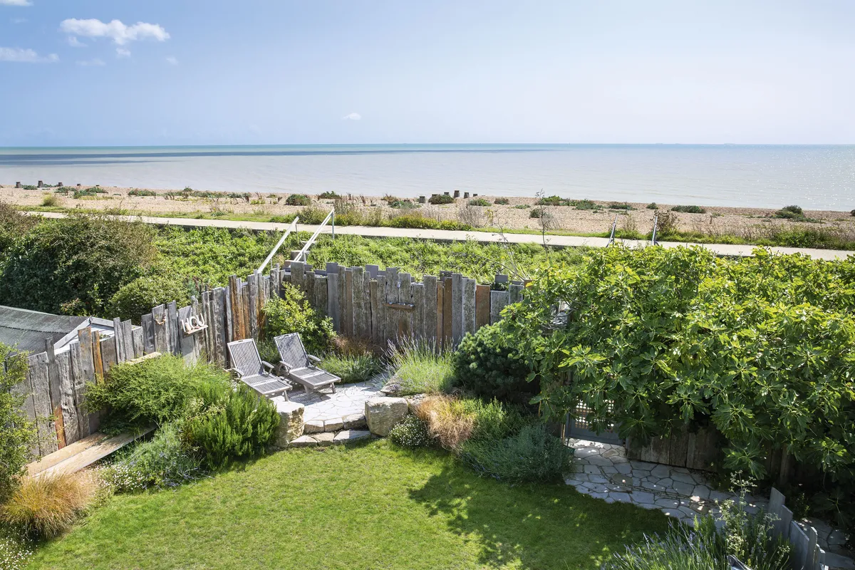 Hunkered down behind the sea wall and surrounded with a weathered timber palisade fence, the garden is protected from the elements with a sunny seating area to the left and shaded corner to the right. A stone path leads to a gate and steps over the sea wall to the beach beyond.