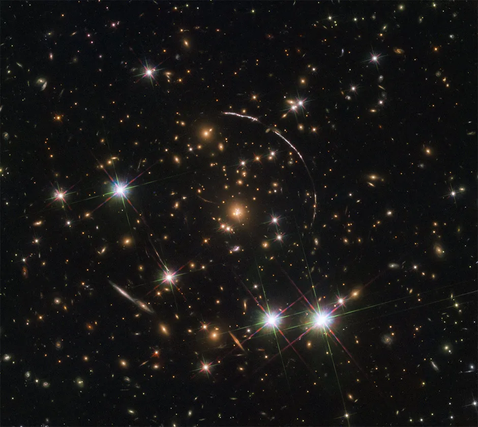 This image, taken by Hubble, is a good example of gravitational lensing. The massive galaxy cluster in the centre of the image is about 4.6 billion light-years away. It is surrounded by four arcs (three in the top right, one in the bottom left). Within these narrow arcs are at least 12 copies of a galaxy nicknamed the Sunburst Arc, located nearly 11 billion light-years away. Its light is being distorted into multiple images by gravitational lensing © ESA/Hubble, NASA, Rivera-Thorsen et al.