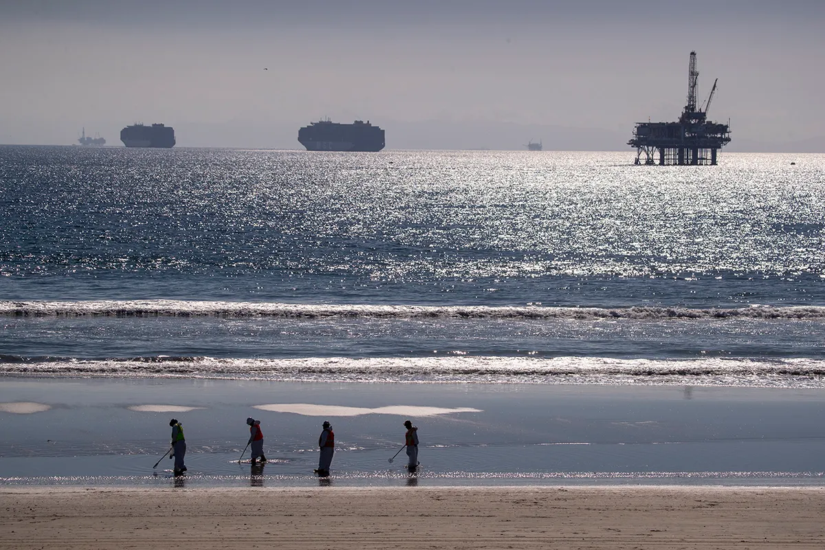Huntington Beach, CA - October 05: Container ships and an oil derrick line the horizon as environmental oil spill cleanup crews search the beach, cleaning up oil chucks from a major oil spill in Huntington Beach Tuesday, Oct. 5, 2021. Environmental cleanup crews are spreading out across Huntington Beach and Newport Beach to cleanup the damage from a major oil spill off the Orange County coast that left crude spoiling beaches, killing fish and birds and threatening local wetlands. The oil slick is believed to have originated from a pipeline leak, pouring 126,000 gallons into the coastal waters and seeping into the Talbert Marsh as lifeguards deployed floating barriers known as booms to try to stop further incursion, said Jennifer Carey, Huntington Beach city spokesperson. At sunrise Sunday, oil was on the sand in some parts of Huntington Beach with slicks visible in the ocean as well. We classify this as a major spill, and it is a high priority to us to mitigate any environmental concerns, Carey said. Its all hands on deck. (Allen J. Schaben / Los Angeles Times via Getty Images)