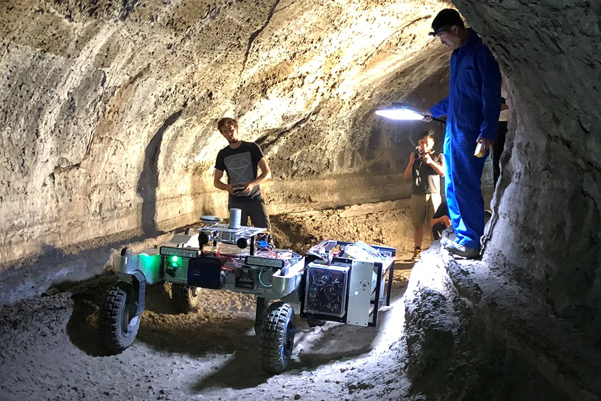 NASA's robotics team drives the test rover, CaveR, into Valentine Cave at Lava Beds National Monument. The science instruments, visible in the box-like structure pointing to one wall of the cave, will begin testing further downstream in the cave. One of the CaveR engineers is perched on a lava ledge, a marker of one of the lava flows in the cave. Photo by NASA/JPL