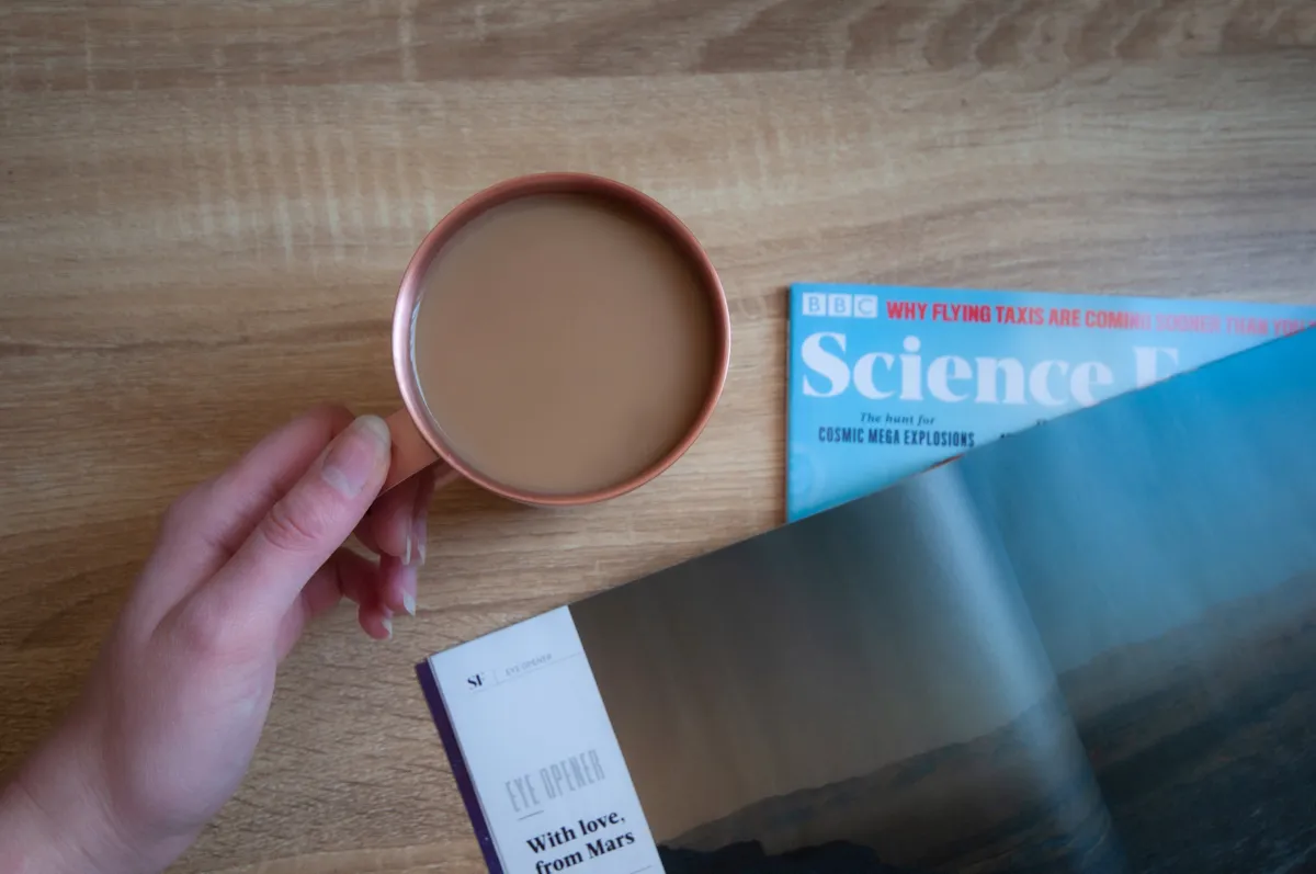 One hand holding a rose gold Ember mug next to an issue of Science Focus magazine