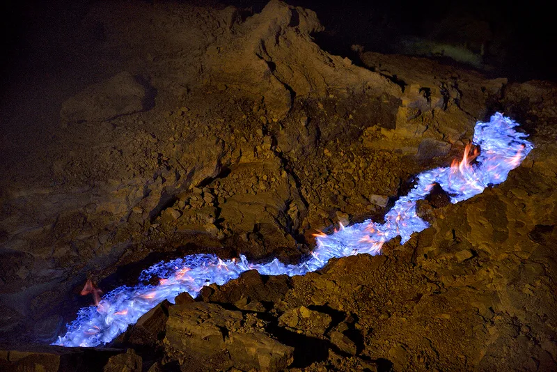 Electric blue lava flows through a a channel in volcanic rock while orange and blue flames flicker from its surface