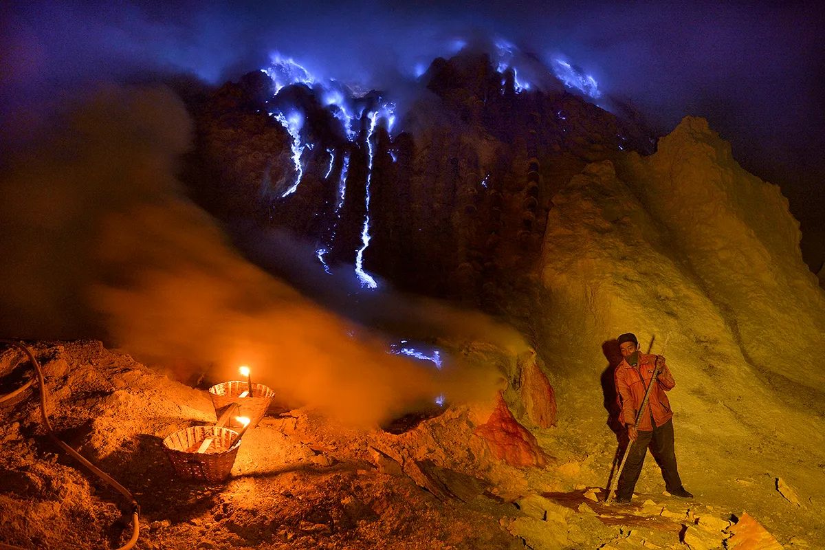 A man collects sulphur at night as blue lava and flames flow and flicker above him in the background