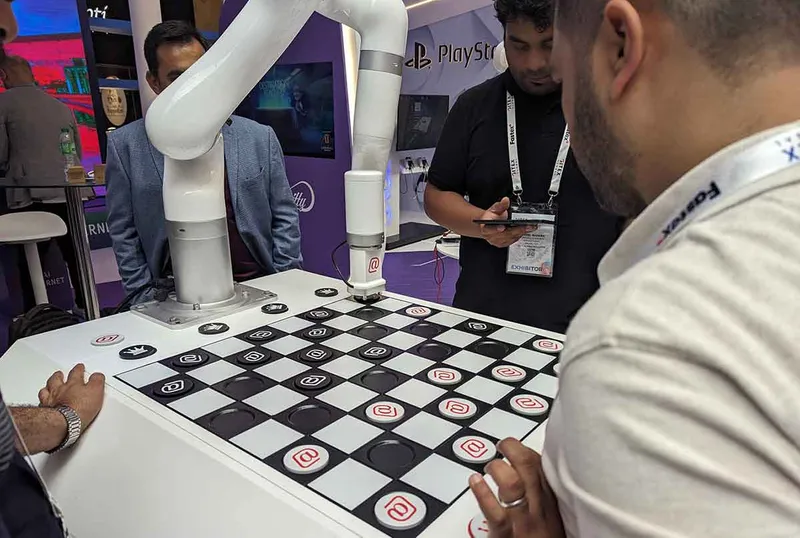 A robot arm sat over a checkers board with a human opponent stood opposite