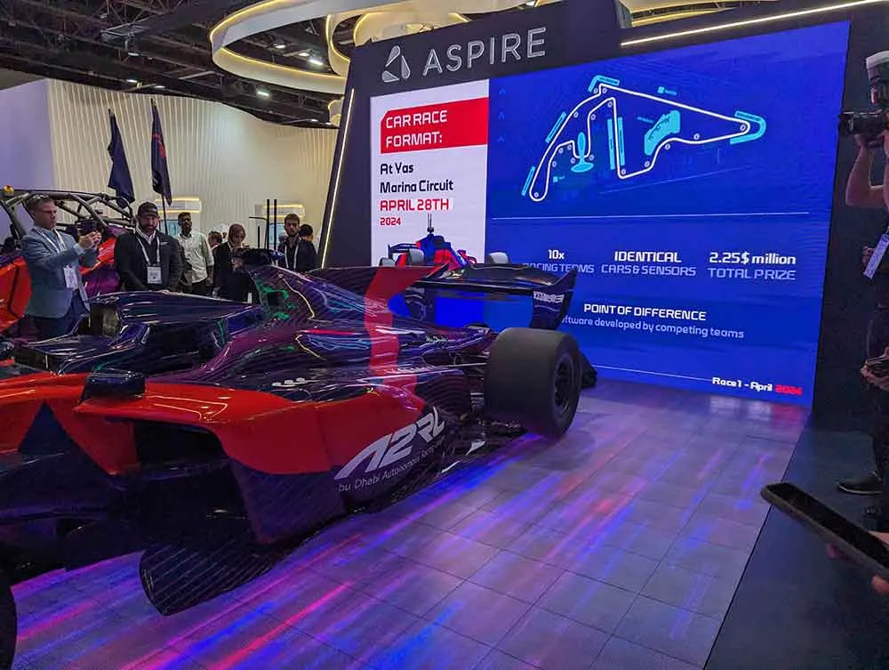 The red and purple autonomous racing car sits on a glowing stage in front of a screen displaying details of the race.