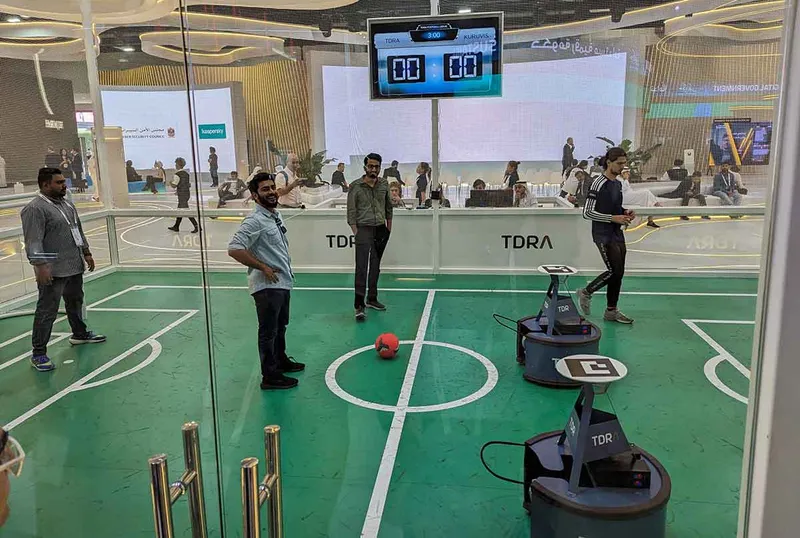 A small football pitch inside a glass room with robots on one side and people on the other.