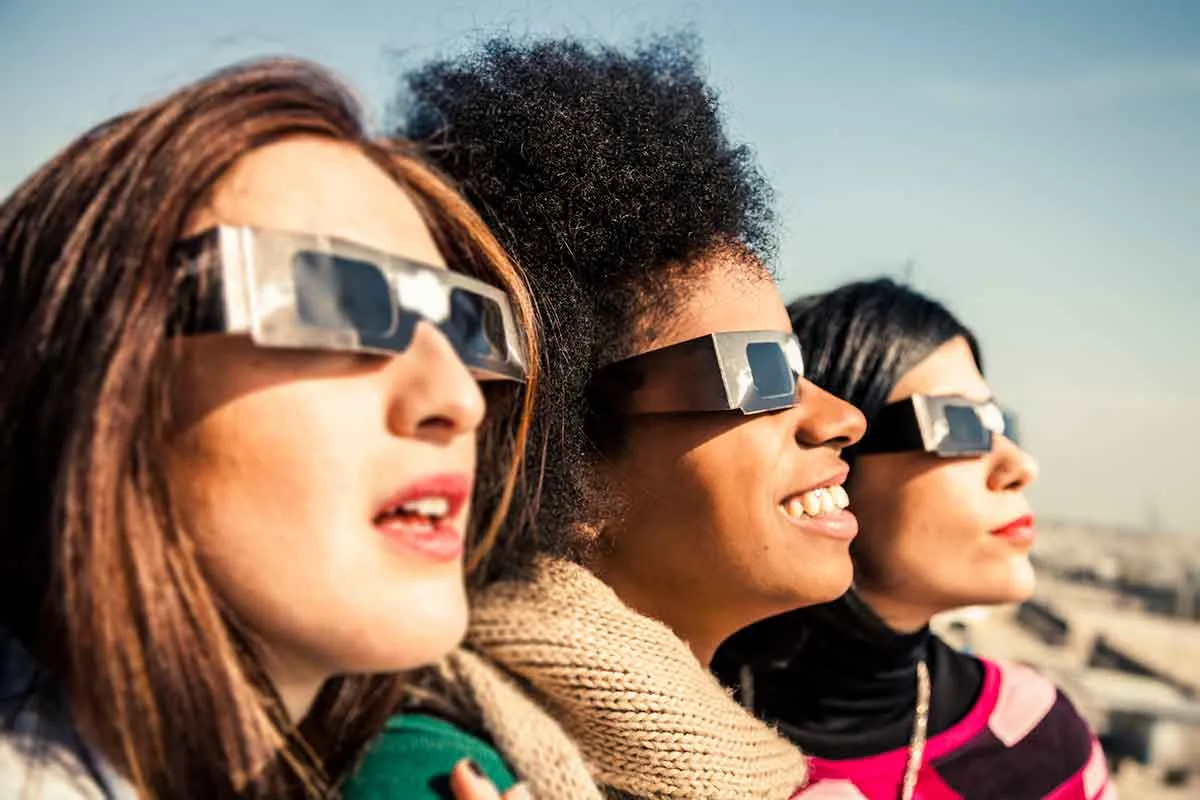 Three women stood in a line wearing eclipse glasses looking at the Sun