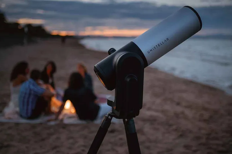 The Unistellar telescope set up on the beach with a ground sat around a BBQ behind.