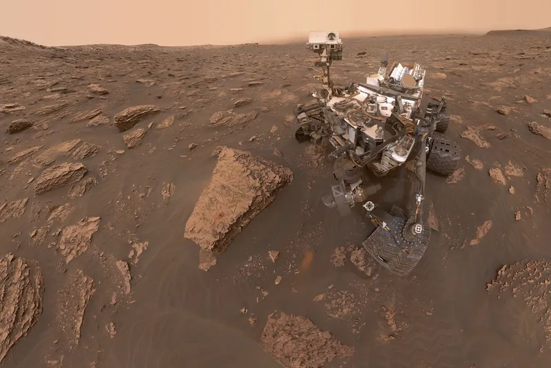 A wheeled rover explores a rusty, rocky planet.
