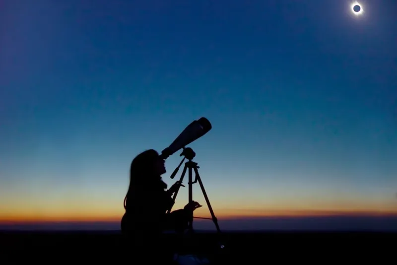 The silhouette of a person observing an eclipse with a telescope. The horizon is reddish in colour, as if it's sunset.