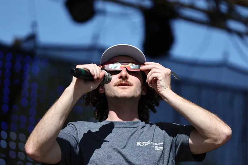 Man holds microphone and wears eclipse glasses.