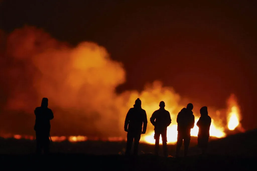 The silhouettes of people watching a fiery eruption in the distance.  