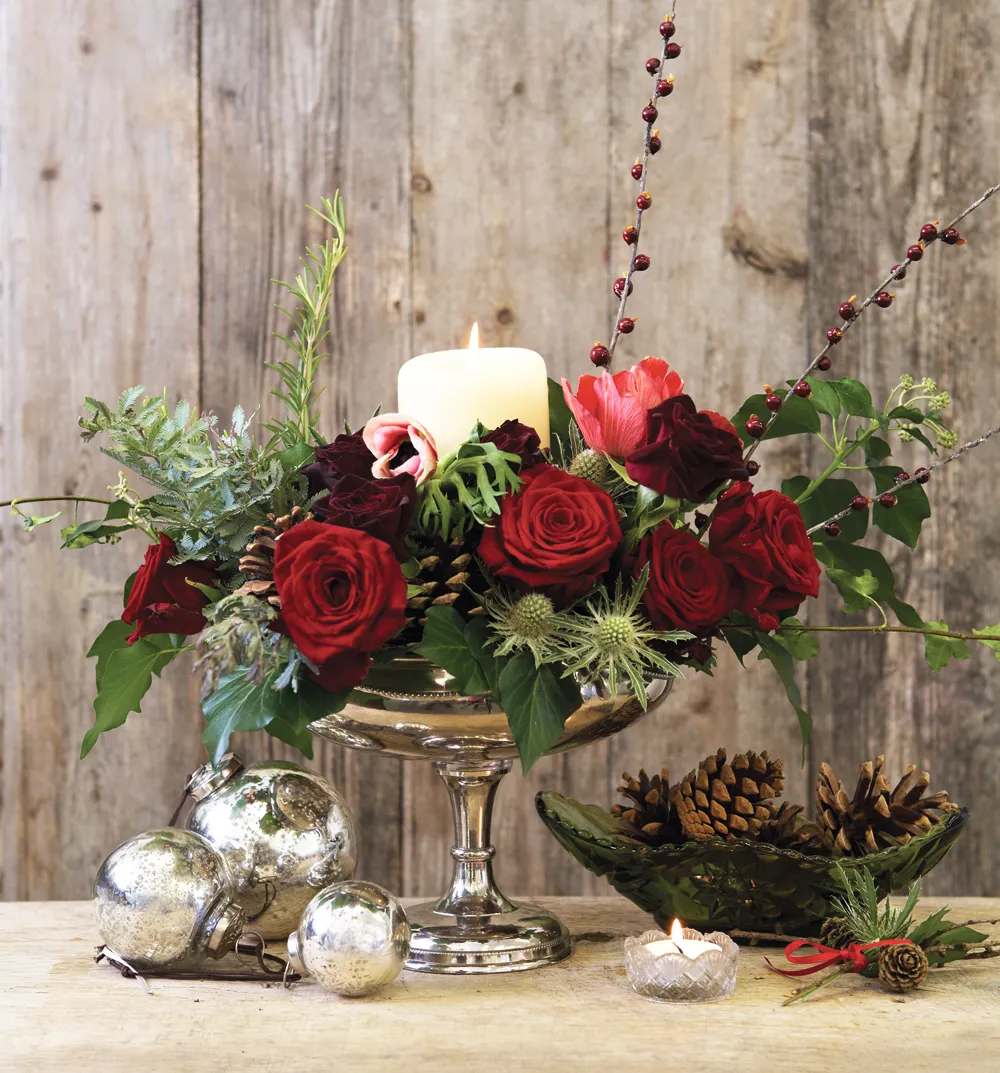 A silver pedestal vase filled with red roses, greenery and a lit pillar candle
