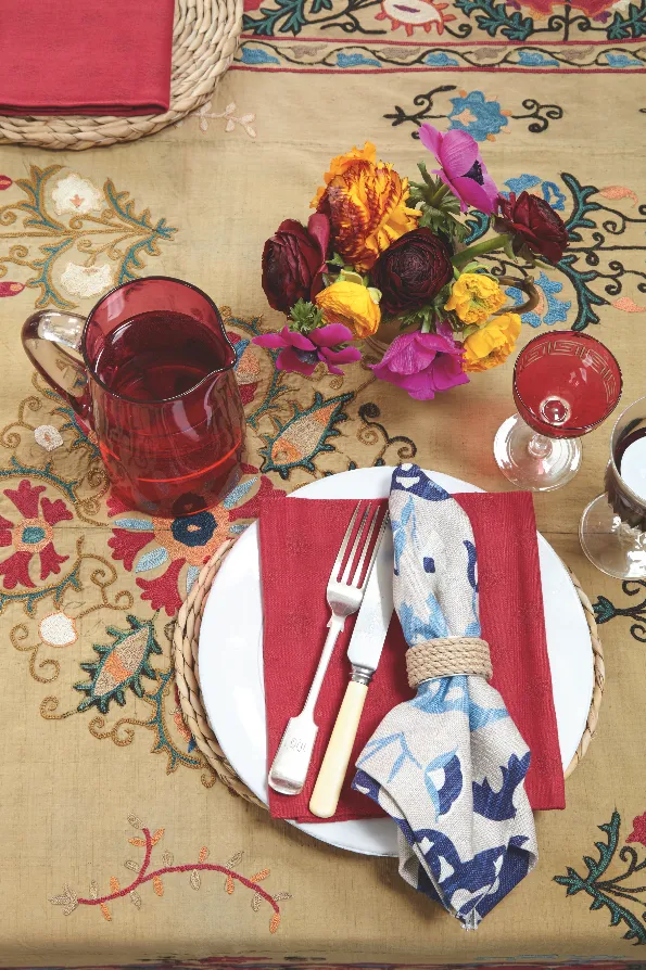 A dining table covered in an antique Suzan with red cranberry glasses, white plates, vintage cutlery and Suzani napkins