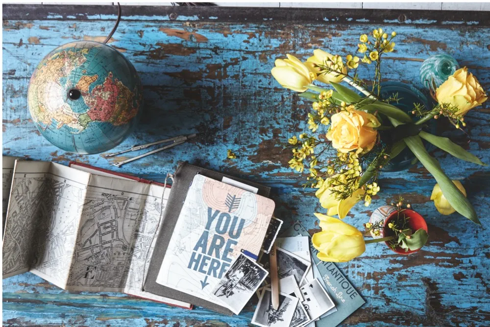 A distressed blue table with an antique globe, map and vase of fresh yellow flowers