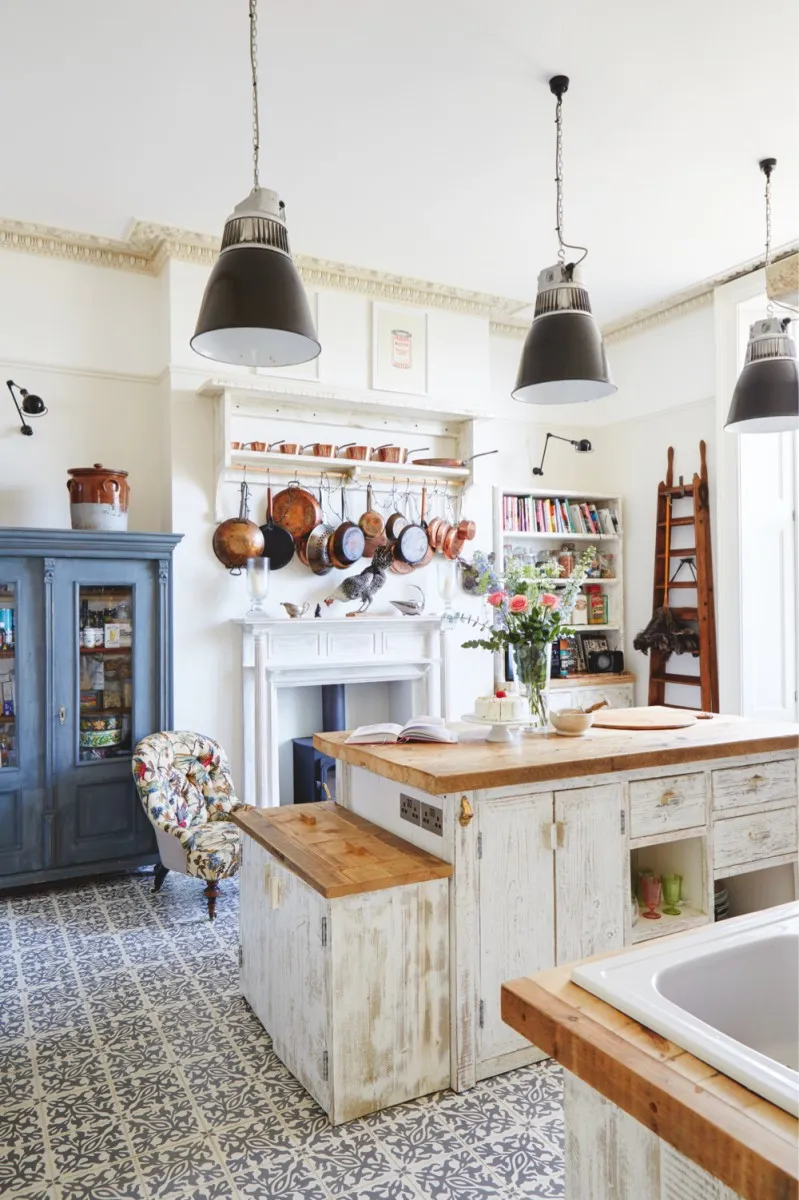 A farmhouse-style white kitchen with blue patterned floor tiles. A collection of vintage copper pans hangs above the fireplace, alongside a painted blue armoire.