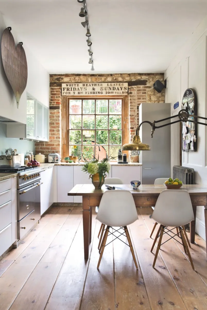 A modern-style kitchen featuring white gloss kitchen units. Four white Eames DSW chairs sit around a farmhouse-style table and a brass vintage lamp hangs overhead.