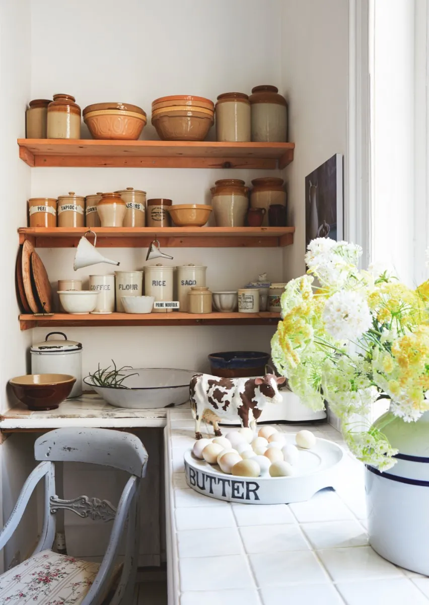 A display of brown pottery and food storage jars in an old-fashioned kitchen. An overflowing vase of gypsophila is in the foreground.