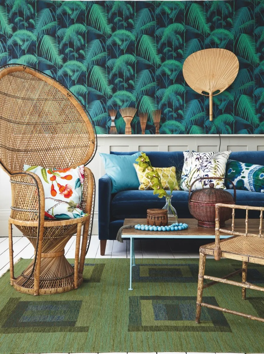 A living room scene with palm-printed wallpaper, a deep blue velvet sofa, a mid-century coffee table and a rattan peacock chair