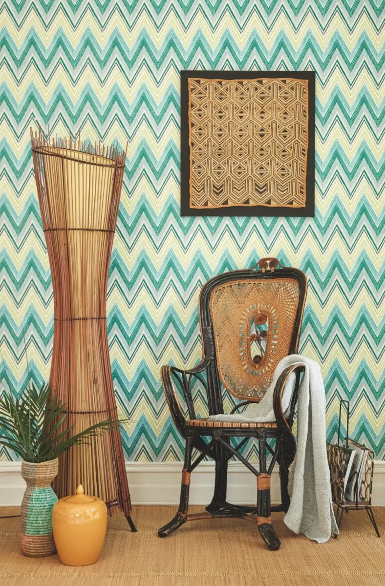 A rattan armchair and tall rattan lamp in front of green and blue zigzag wallpaper