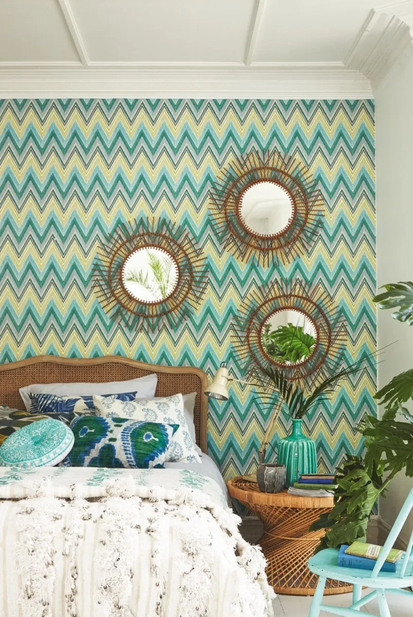 A double bed with a woven rattan headboard against green and blue zigzag wallpaper with three rattan mirrors