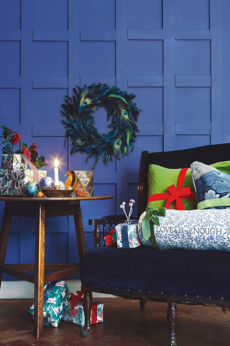 Blue velvet chair next to an arts and crafts table against a vibrant blue background Presents wrapped in Emily Burningham wrapping paper, ribbons tied around patterned cushions, art deco baubles, Morris & Co accessories and arts and crafts antiques