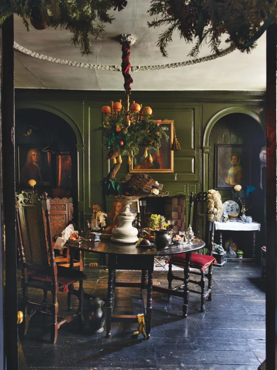 The dining room in Dennis Severs' House with a dark wood table, dark green walls, oil paintings and a chandelier adorned with greenery and oranges