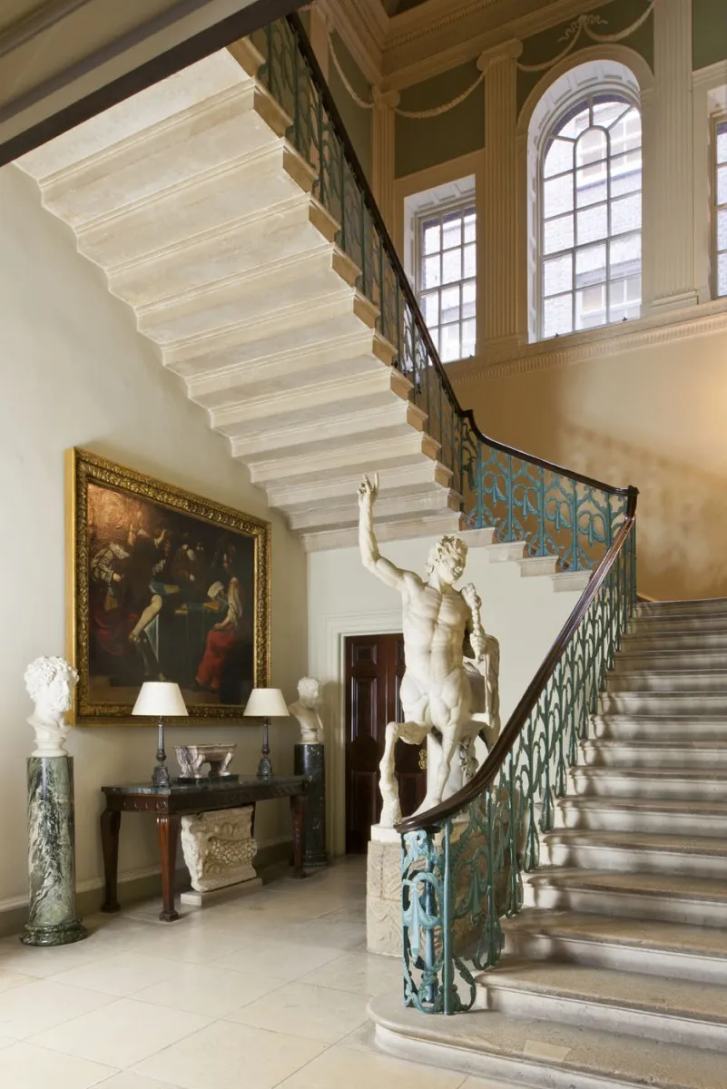 The grand staircase hall in Spencer House, featuring a sweeping stone staircase with an Grecian sculpture at the bottom.