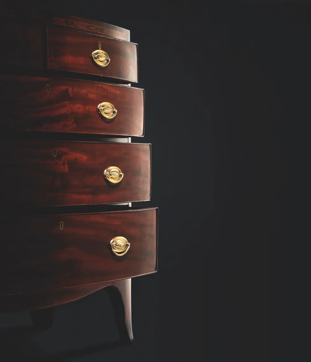 A side-on shot of a Georgian bow-fronted chest of drawers against a black background