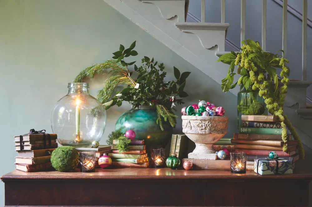 A display of vintage books, antique baubles, festive foliage and candles on a console table in a festive hallway.